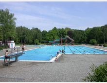 Ein Freibad mit einem großen rechteckigen Schwimmbecken, umgeben von einer gepflasterten Fläche und Rasenflächen im Hintergrund. Zu sehen ist ein Sprungbrett, eine Wasserrutsche und Bäume, die das Gelände einrahmen.