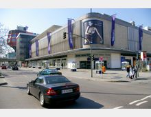 Straßenszene vor einem Kaufhaus mit moderner Fassade und der Aufschrift „Wertheim“. Autos fahren auf der Straße, und Menschen gehen auf dem Bürgersteig; im Hintergrund ist ein markantes rotes Gebäude mit auffälliger Architektur sichtbar.