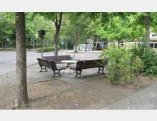 Sitzbereich im Freien mit drei Holzbänken um einen runden Tisch, umgeben von Bäumen und Büschen. Im Hintergrund sind eine Straßenkreuzung mit Ampeln, Verkehrsschildern und ein Schulgebäude zu sehen.