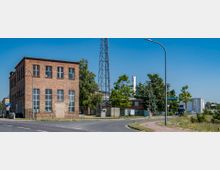 Ziegelgebäude mit großen Fenstern auf der linken Seite, umgeben von einem Industriegelände mit Bäumen und einem hohen Stahlgittermast im Hintergrund; im Vordergrund eine Straße mit Laternen und einem Weg.
