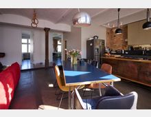 Offener Wohnraum mit moderner Küche und Essbereich, ausgestattet mit einem langen blauen Tisch und bunten Stühlen. Im Hintergrund sind sichtbare Backsteinwände, eine freistehende Stützsäule, große Fenster und Geräte wie ein silberner Kühlschrank zu sehen.