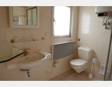 Kleines Badezimmer mit beigefarbenen Fliesen, einem Waschbecken mit Glasablage, einem Spiegel, einer Toilette, einem Heizkörper unter einem Fenster mit Vorhang und einer Duschkabine am rechten Rand. Ein Warmwasserboiler ist an der Wand montiert.