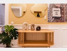 Innenraumansicht mit einer gelben Wand, darauf zwei organisch geformte Spiegel und ein gerahmtes Kunstwerk. Im Vordergrund steht ein Holzregal mit drei abstrakten Keramikfiguren und daneben eine grüne Monstera-Pflanze.