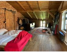 Dachgeschosszimmer mit roten Ziegelwänden, sichtbar offenen Holzbalken und Holzdecke. Der Raum enthält ein Bett mit weißer Bettwäsche und einer roten Decke, einen Teppich, eine Kleiderstange mit Kleidung, ein Holzschreibtisch am Fenster und Holzböden.