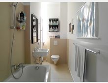 Modernes Badezimmer mit Badewanne, Wand-Duschkopf, kleinem Waschbecken und Toilette. Wände sind teilweise beige gefliest, mit Handtuchhalter, einem Spiegel, Regalen mit Pflegeprodukten und einem Wandbild an der schlichten weißen Wand.