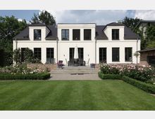 Weiße Villa mit dunklem Schieferdach und mehreren großen Fenstern, umgeben von einem gepflegten Garten mit Rasenfläche, Blumenkästen und Rosenbüschen; eine Terrasse mit Gartenmöbeln und Sonnenschirm befindet sich zentral vor dem Gebäude.