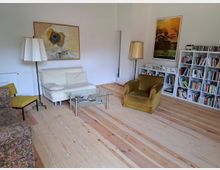 Wohnzimmer mit Holzdielenboden, heller Wandfarbe und verschiedenen Möbelstücken, darunter ein weißes Sofa, ein gelber Sessel und ein floral gemusterter Sessel. An der Wand hängen zwei gerahmte Bilder, daneben stehen eine Bücherwand und Stehlampen. Ein Glastisch steht in der Mitte des Raumes.