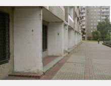 Reihen von Plattenbau-Wohnhäusern mit grauen Fassaden und Balkonen, daneben ein gepflasterter Gehweg mit vereinzelten roten Klinkersteinen. Im Hintergrund sind Bäume und eine Person zu Fuß zu sehen.