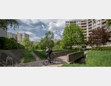 Ein Wohngebiet mit mehrstöckigen Wohngebäuden im Hintergrund, umgeben von grünen Bäumen und Rasenflächen. Im Vordergrund fährt eine Person mit einem Fahrrad über eine kleine Brücke aus Beton.