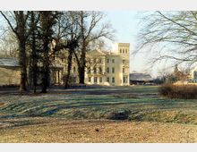 Großes, cremefarbenes Herrenhaus mit historischen Architekturelementen, darunter Säulen und ein Turm, umgeben von Bäumen und einer weitläufigen Wiesenfläche in einer parkähnlichen Landschaft. Im Hintergrund sind weitere kleinere Gebäude sichtbar.