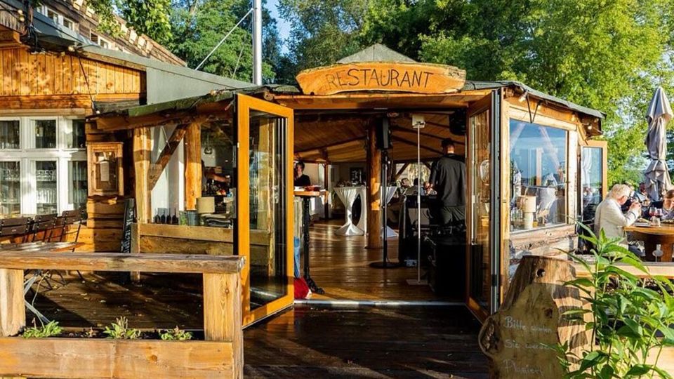 Holzrestaurant mit offenem Eingang und überdachtem Innenbereich, umgeben von einer Terrasse mit Holzmöbeln. Im Hintergrund befinden sich Bäume und Sonnenschirme, die eine Außenbewirtung andeuten.