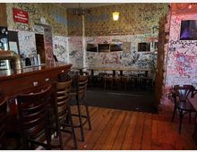 Innenraum einer Kneipe mit dunkler Holztheke und Barhockern auf Holzdielenboden. Die Wände und die Decke sind dicht mit Graffiti und Kritzeleien bedeckt, dazu einfache Tische und Stühle sowie eine kleine erhöhte Sitzecke an der Rückwand.