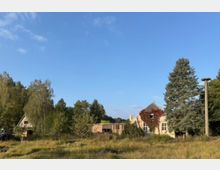 Eine ländliche Szene mit einem Wiesenbereich im Vordergrund, einem mit Efeu bewachsenen Haus rechts und einem Holzgebäude links, umgeben von Bäumen und Wald. Neben dem Haus steht ein hoher Betonpfahl mit einer Plattform an der Spitze unter klarem blauem Himmel.
