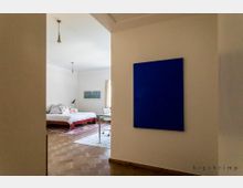 Flur mit Blick in ein Schlafzimmer. Der Raum hat helle Wände, einen Holzboden, ein großes Bett mit bunter Bettwäsche und Teppich sowie ein Fenster mit Vorhang. An der Wand im Flur hängt ein großes, minimalistisches, blaues Kunstwerk.