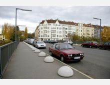Eine städtische Straßenszene mit geparkten Autos entlang einer Brücke, flankiert von breiten Gehwegen mit runden Betonkugeln als Begrenzung. Im Hintergrund sind mehrstöckige Wohnhäuser mit hellen Fassaden und Balkonen, sowie herbstliche Bäume zu sehen.