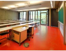 Klassenzimmer mit roten Böden, weißen Tischen und schwarzen Stühlen, einer Lehrerpult vorne links sowie einer großen Tafel an der rechten Wand. Große Fenster und Glastüren in der Rückwand lassen Tageslicht herein und bieten Ausblick auf grüne Bäume.