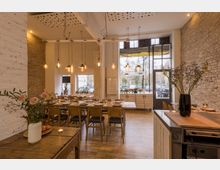 Innenraum eines modernen Restaurants mit hohen Decken, freiliegenden Backsteinwänden und großen Fenstern, die Tageslicht hereinlassen. Ein großer Holztisch ist mit Geschirr und Gläsern für ein Essen gedeckt, und hängende Glühbirnen dienen als Beleuchtung.