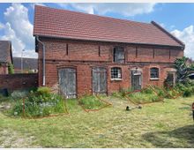 Backsteinbau mit rotem Ziegeldach in ländlicher Umgebung, erkennbar an mehreren Holztoren, kleinen Fenstern und verwilderter Vegetation im Vorgarten. Im Vordergrund ein Rasenbereich sowie vereinzelt Gartengeräte und Pflanzenbeete.