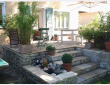 Steinterrasse mit Treppenstufen, bepflanzten Blumentöpfen und einem kleinen Wasserbecken mit Steinen, angrenzend an ein Haus mit cremefarbener Fassade, offenem Fensterladen und Gartenmöbeln unter einem Sonnenschirm.