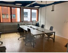 Besprechungsraum mit Glastrennwand, hellem Holzboden und weiß gestrichener Ziegelwand. Der Raum ist mit einem langen weißen Tisch, mehreren schwarzen Bürostühlen und großen Fenstern mit Blick nach draußen ausgestattet.