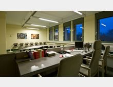 Ein Büroraum mit mehreren Schreibtischen und gepolsterten Stühlen. Im Vordergrund stehen Arbeitsplätze mit Computern und Dokumenten, im Hintergrund befinden sich eine Reihe von Besucherstühlen, große Fenster und Wanddekorationen in Form von Bildern.