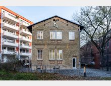 Fassade eines historischen Backsteingebäudes mit giebelförmigem Dach und weißen Fensterrahmen, umgeben von modernen Wohnhäusern und Bäumen. Im Vordergrund ein gepflasterter Weg und herbstliches Laub, rechts ein dekorativer Zaun.