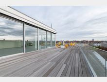 Dachterrasse mit Holzboden und Glasgeländer, ausgestattet mit gelben Liegestühlen und einem weißen Tisch, angrenzend an eine moderne Glasfassade; im Hintergrund Blick auf umliegende städtische Gebäude unter bewölktem Himmel.