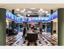 Foyer eines modernen Gebäudes mit geometrischem Bodenmuster, einer runden Empfangstheke mit Blumenarrangement und gläsernen Drehtüren. Im oberen Bereich befindet sich ein leuchtendes Band mit farbenfrohen Cartoon-Illustrationen.