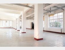 Heller, leerer Industrie- oder Loft-Innenraum mit glattem Betonboden und mehreren weißen Tragstützen mit roten Sockeln. Große Sprossenfenster lassen viel Tageslicht herein; an der Decke sind sichtbare Rohrleitungen und Kabeltrassen.