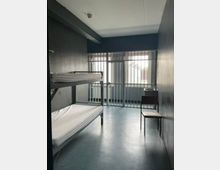 Schmale, karge Zelle mit grauen Wänden, einem Metall-Etagenbett mit dünnen Matratzen und einem einzelnen Stuhl. Am Ende des Raums befindet sich eine vergitterte Tür vor einem Fenster, durch das Tageslicht einfällt.