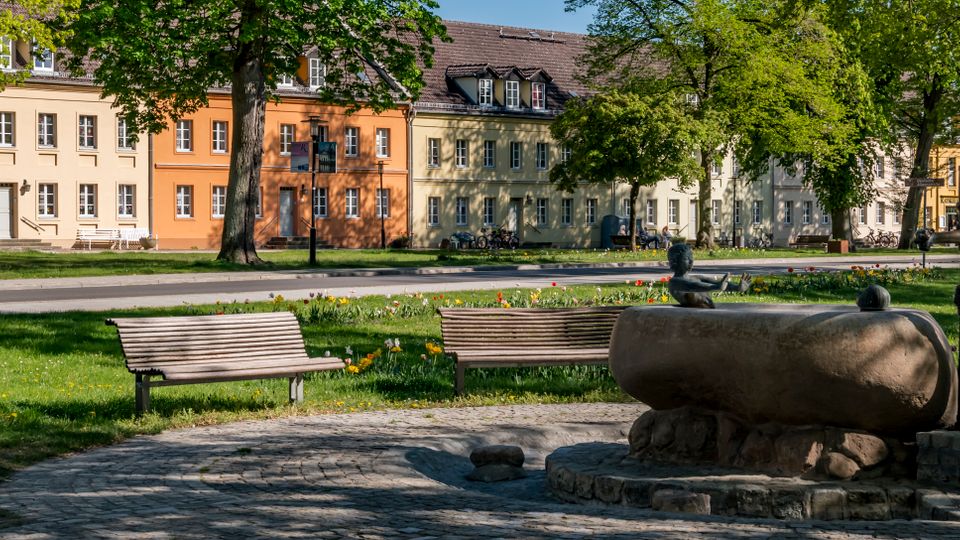 Tree-lined park plaza with wooden benches and a cobblestone area featuring a stone trough and old hand water pump in the foreground. Across a road, a row of pastel-colored historic buildings with many windows and a tiled roof forms the background.