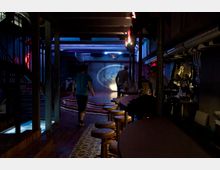Dunkle Bar mit Holz- und Metall-Elementen, einer Theke mit Barhockern auf der rechten Seite und einem erhöhten, beleuchteten Podest im Hintergrund. Gedämpfte bunte Beleuchtung und Dekorationen schaffen eine Club-Atmosphäre.