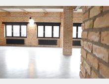 Loftartiger Raum mit Backsteinwänden, großen schwarzen Fenstern und dunklen Heizkörpern unter den Fenstern. Der Boden ist aus glänzendem Beton, und Betonträger sind an der Decke sichtbar.