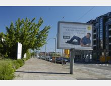 Straßenszene mit einer Gehwegfläche, geparkten Autos am Straßenrand und einer großen Werbetafel im Vordergrund, die ein Mobilfunkangebot bewirbt; im Hintergrund sind moderne Gebäude und ein Baum auf der linken Seite sichtbar.