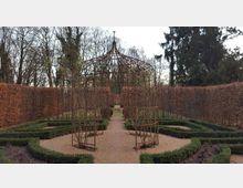 Metallpavillon in einem symmetrischen Garten mit Kieswegen und niedrigen Buchsbaumhecken, umgeben von hohen, kahlen Hecken und Bäumen im Hintergrund.