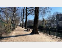 Gepflegter Fußweg entlang eines Kanals mit hohen, laublosen Bäumen und Metallgeländer auf der rechten Seite. Links des Weges befindet sich ein Drahtzaun mit vereinzeltem Buschwerk, und im Hintergrund sind Wohnhäuser zu sehen.