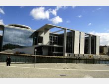 Modernes Verwaltungsgebäude mit großer Glasfassade und geometrischen Betonstrukturen, gelegen an einem Wasserkanal. Im Vordergrund eine gepflasterte Promenade mit Geländer am Ufer.