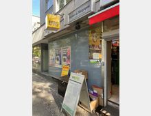 Außenansicht eines kleinen Zeitungskiosks in einer belebten städtischen Umgebung. Zu sehen sind ein BVG-Schild, Werbeschilder für Lotto und Zeitungen sowie ein aufgestelltes Schild mit Postpreisinformationen vor dem Eingang.