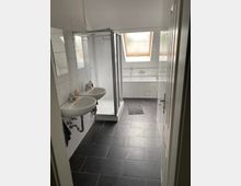 Modernes Badezimmer mit dunklem Fliesenboden, zwei Waschbecken, einer verglasten Duschkabine und einer Badewanne unter einem Dachfenster. Die Wände sind weiß gefliest, und Tageslicht fällt durch das Fenster ein.