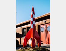 Eine rote Modellrakete mit weißen Karos steht draußen vor einem Gebäude mit Holzfassade und roten Türen. Der Himmel ist klar und blau.