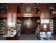 Historische Bibliothek mit dunklen Holzregalen, gefüllt mit Büchern, und detaillierten Verzierungen. In der Mitte ein großer Holzpfeiler mit einer analogen Uhr darüber und einer Holztür im Hintergrund.