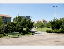 Städtischer Park mit einer gepflasterten Fläche im Vordergrund, einer Holzbank und Grünanlagen mit Bäumen und Sträuchern. Im Hintergrund sind moderne und historische Gebäude sowie ein klarer blauer Himmel sichtbar.