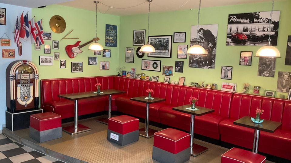 Innenraum eines Retro-Diners mit langen roten Kunstleder-Sitzbänken, kleinen quadratischen Tischen und passenden Hockern auf einem schwarz-weiß karierten Boden. An der hellgrünen Wand hängen zahlreiche gerahmte Fotos und Musikdekorationen, daneben steht eine Jukebox; mehrere Hängelampen beleuchten die Sitzreihe.