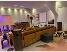 Wohnzimmer mit modernem, gemütlichem Design: Ein senfgelbes Sofa steht im Mittelpunkt auf einem Kuhfellteppich, daneben Holzmöbel und eine Stehlampe. Im Hintergrund befinden sich ein Sideboard mit Plattenspielern, Lautsprecher und dekorative Bilder an der Wand. Ein Hund liegt auf dem Teppich.