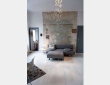 Wohnzimmer mit hell gestrichenen Holzböden, einer grauen Couch und einem dazugehörigen Hocker in der Mitte. Eine raue Wand mit sichtbaren Putz- und Farbresten dient als gestalterisches Element, dekoriert mit kleinen Bildern; moderne Möbel und eine hängende Lichtinstallation betonen das minimalistische Design.