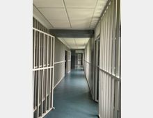Langer Flur in einem Haft- oder Gefängnistrakt mit blauem Boden, abgehängter Decke und mehreren offenen Gittertüren entlang der Seiten. Die Sicht führt zu weiteren Gitterabschnitten und Türen am Ende des Korridors.