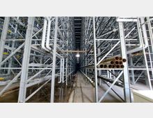 Innenansicht eines hohen automatisierten Lagersystems mit zahlreichen Stahlregalen, die sich auf beiden Seiten in die Tiefe erstrecken. Sichtbare Rohre und Stahlträger prägen die industrielle Umgebung, im Vordergrund liegen dicke Metallrohre auf den Regalen.