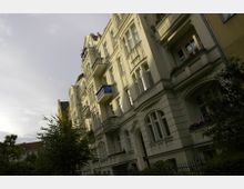 Mehrstöckiges Wohngebäude im Altbaustil mit hellen Fassaden und dekorativen Details wie Rundbogenfenstern und Balkonen; im Vordergrund Bäume und ein schmiedeeiserner Zaun. Bewölkter Himmel im Hintergrund.