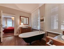 Helles Badezimmer mit freistehender Badewanne auf Holzfußboden, einem Waschtisch mit Spiegel und Lampe sowie großen Fenstern mit blickdichten Jalousien. Im Hintergrund ist ein angrenzendes Schlafzimmer mit roter Bettdecke und Vorhängen sichtbar.