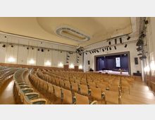 Ein moderner Veranstaltungssaal mit gebogenen Sitzreihen aus Holzstühlen, einer Bühne mit schwarzem Vorhang, einem Flügel und einem Rednerpult. An der Decke befinden sich ein dekoratives Oval-Element und Bühnenscheinwerfer, die an Metallkonstruktionen befestigt sind.